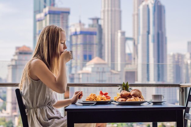 Une jeune femme prend son petit déjeuner sur le balcon. Table de petit-déjeuner avec café, fruits et pain croissant sur un balcon dans le contexte de la grande ville