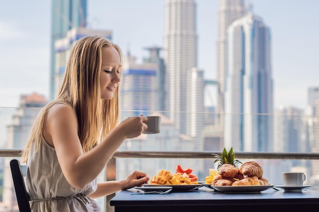 Une jeune femme prend son petit déjeuner sur le balcon. Table de petit-déjeuner avec café, fruits et pain croissant sur un balcon dans le contexte de la grande ville