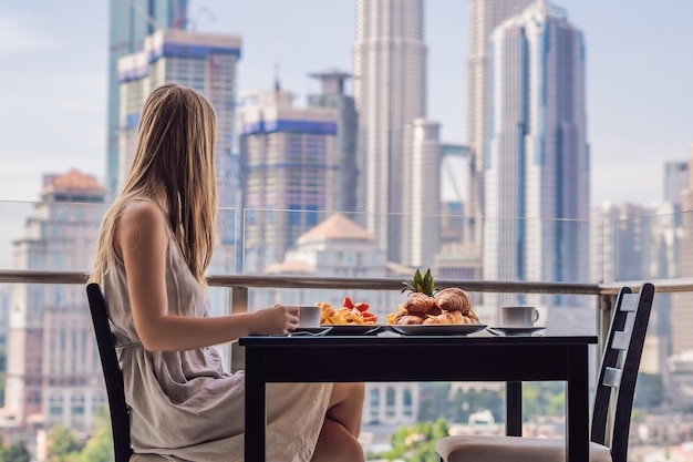 Une jeune femme prend son petit-déjeuner sur le balcon Table de petit-déjeuner avec café, fruits et pain croissant sur un balcon sur fond de grande ville