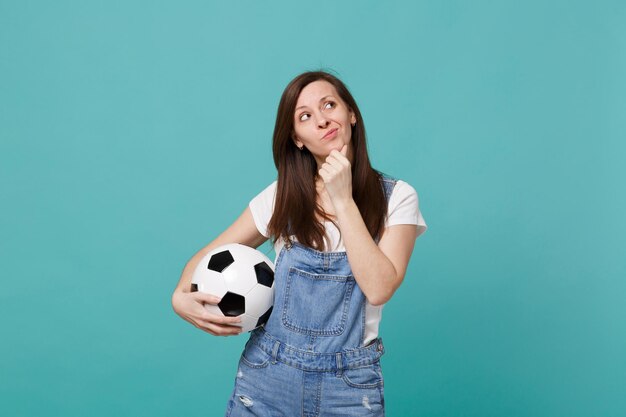 Une jeune femme préoccupée par un fan de football soutient l'équipe préférée avec un ballon de football en levant, place la main sur le menton isolé sur fond bleu turquoise. Émotions des gens, concept de loisirs sportifs en famille.