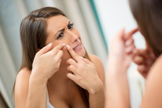 La jeune femme a des problèmes de peau sur le visage. Elle regarde un bouton sur sa joue dans le miroir et fronce les sourcils.