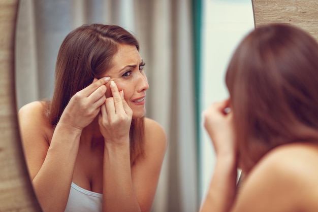 La jeune femme a des problèmes de peau sur le visage. Elle regarde un bouton sur sa joue dans le miroir et fronce les sourcils.