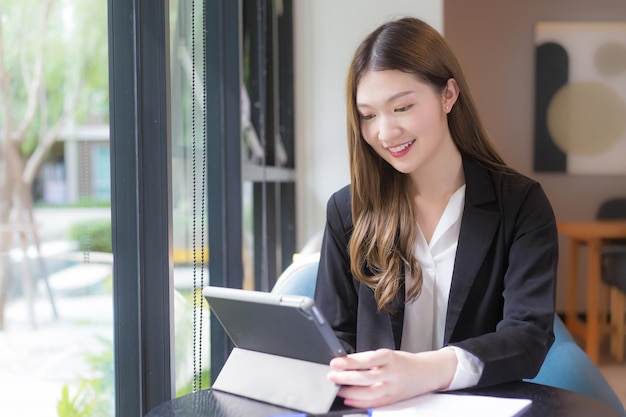 Une jeune femme professionnelle asiatique en costume noir travaille sur une tablette en souriant