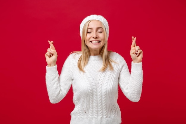 Photo jeune femme en pull, chapeau isolé sur fond rouge. concept de saison froide de mode de vie sain. maquette de l'espace de copie. en attendant un moment spécial, en gardant les doigts croisés, les yeux fermés, en faisant un vœu.