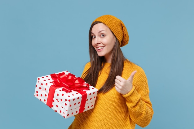 Jeune femme en pull et chapeau posant isolé sur fond bleu. Concept d'anniversaire de la Saint-Valentin, de la femme, de la journée. Maquette de l'espace de copie. Tenez une boîte cadeau rouge blanche avec un noeud de ruban cadeau montrant le pouce vers le haut.