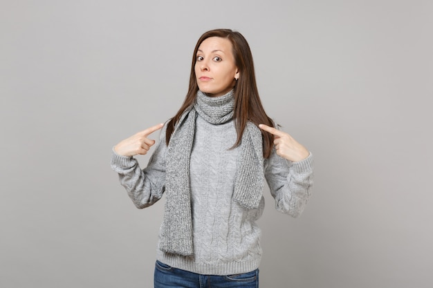 Jeune femme en pull gris, écharpe pointant l'index sur elle-même isolée sur fond gris en studio. Mode de vie sain, émotions sincères, concept de saison froide. Maquette de l'espace de copie.
