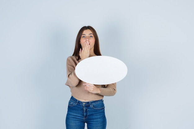 Jeune femme en pull, jeans couvrant la bouche avec la main, tenant une bulle de papier et l'air choqué, vue de face.