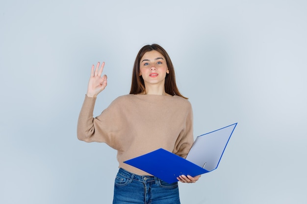 Jeune femme en pull, jeans montrant un geste correct, tenant un dossier et l'air heureux, vue de face.