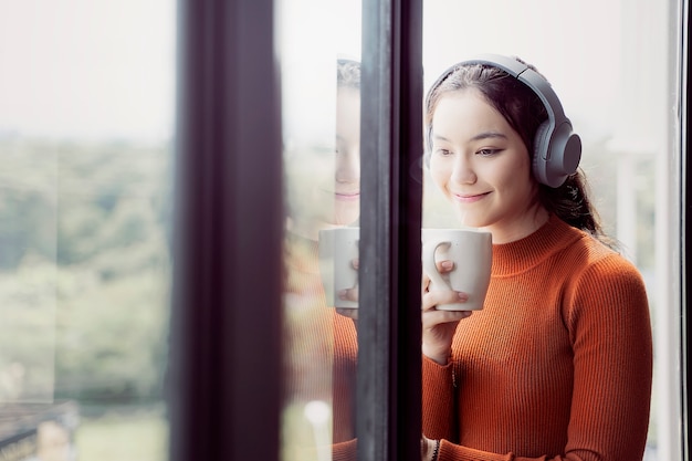 Jeune femme en pull orange écoute musice et tenant la tasse en se tenant debout près de la fenêtre.