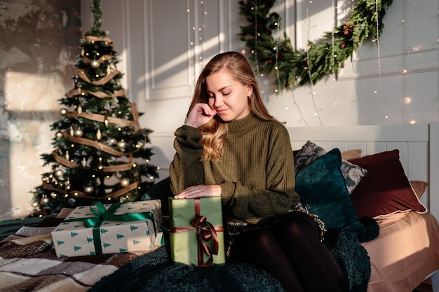 Une jeune femme en pull ouvre des cadeaux de Noël dans une chambre décorée pour Noël sur fond d'arbre de Noël.