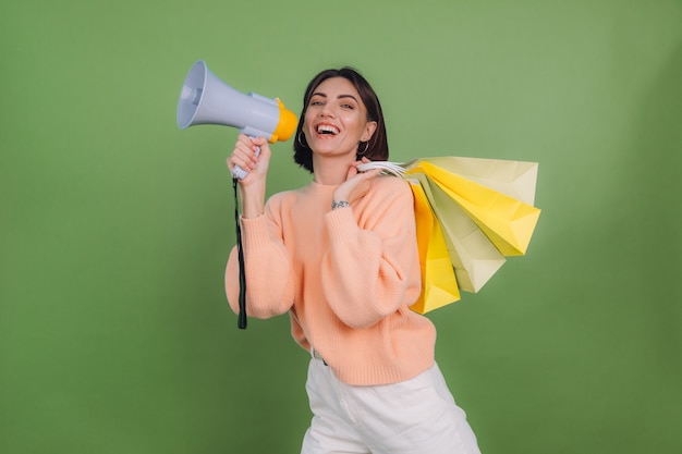 Jeune Femme En Pull Pêche Décontracté Isolé Sur Un Mur De Couleur Vert Olive Cri Dans Un Mégaphone Tenant Des Sacs à Provisions, Annonce Des Remises Promotion De Vente