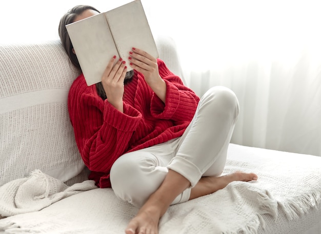 Jeune femme en pull rouge sur le canapé à la maison avec un livre dans les mains.