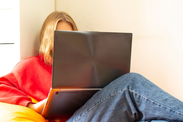 Une jeune femme en pull rouge est assise sur un sac de haricots orange dans la chambre et travaille à la maison avec un ordinateur portable