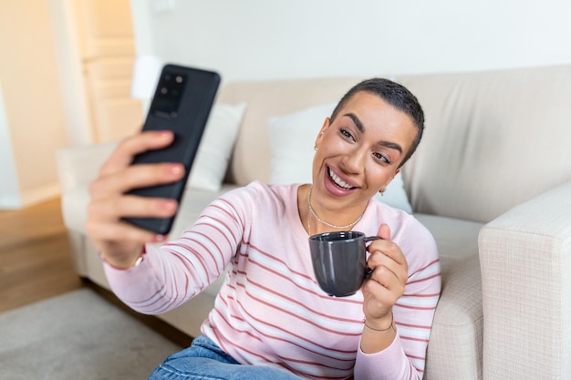 Une jeune femme en pull avec une tasse dans les mains regarde le téléphone assise sur le canapé