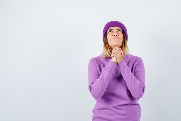 Jeune femme en pull violet, bonnet tenant les mains jointes dans un geste de prière et l'air plein d'espoir, vue de face.
