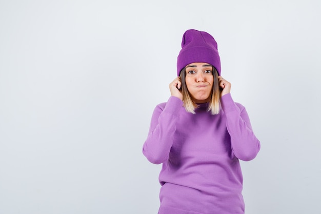 Jeune Femme En Pull Violet, Bonnet Tirant Vers Le Bas Ses Lobes D'oreilles Tout En Soufflant Les Joues Et En Ayant L'air Drôle, Vue De Face.