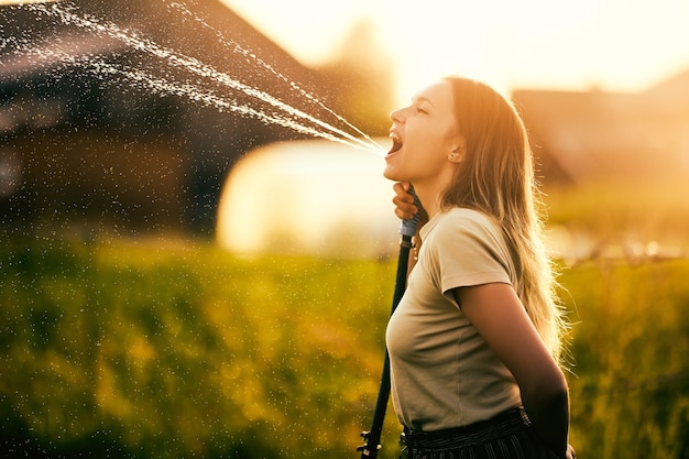 Une jeune femme pulvérise de l'eau du tuyau de jardin pour arroser les plantes tout en s'amusant.