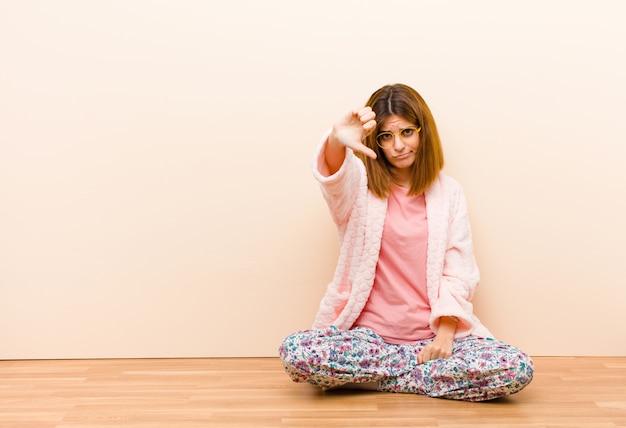 Jeune femme en pyjama assis à la maison se sentant en colère, en colère, ennuyée, déçue ou contrariée, montrant les pouces vers le bas avec un air sérieux