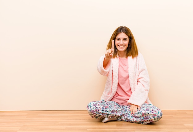 Jeune femme en pyjama assis à la maison en souriant et amical, montrant le numéro un ou le premier avec la main en avant, comptant à rebours