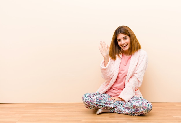 Jeune femme en pyjama assis à la maison en souriant joyeusement et joyeusement en agitant la main pour vous saluer ou vous dire au revoir