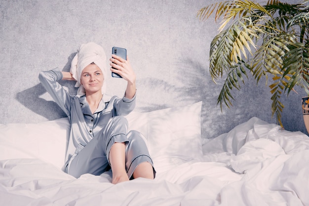 Jeune femme en pyjama et une serviette sur la tête est assise dans son lit et regarde son téléphone