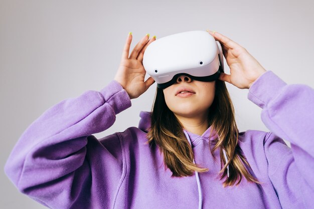 Jeune femme de race blanche à l'aide de casque VR, toucher des lunettes et levant en réalité virtuelle sur fond blanc.