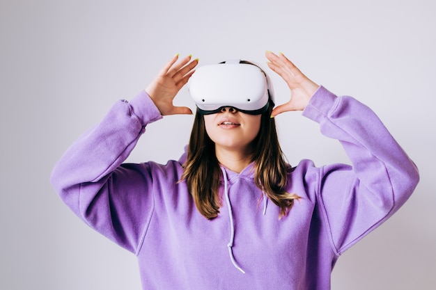 Jeune femme de race blanche à l'aide de casque VR, toucher des lunettes et levant en réalité virtuelle sur fond blanc.