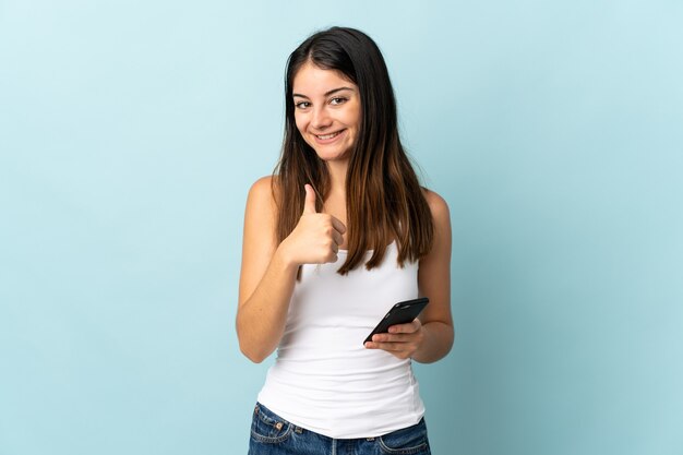 Jeune femme de race blanche à l'aide de téléphone mobile isolé sur bleu donnant un geste de pouce en l'air