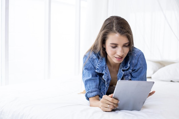 Jeune femme de race blanche allongée accroupie sur le lit jouant socialement en ligne dans une tablette avec plaisir dans la chambre à la maison seul tout en se relaxant.