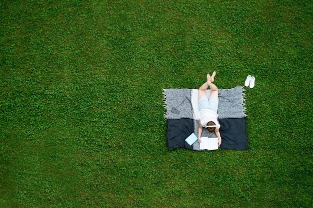 Jeune femme de race blanche allongée sur la couverture sur la pelouse verte et lire un livre