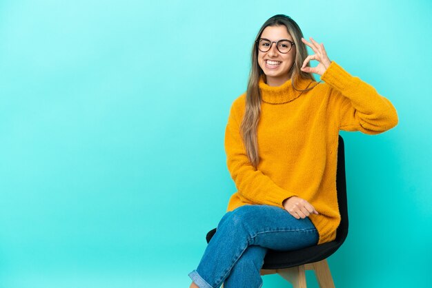 Jeune femme de race blanche assise sur une chaise isolée sur fond bleu montrant signe ok avec les doigts