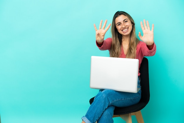 Jeune femme de race blanche assise sur une chaise avec son pc isolé sur fond bleu en comptant neuf avec les doigts