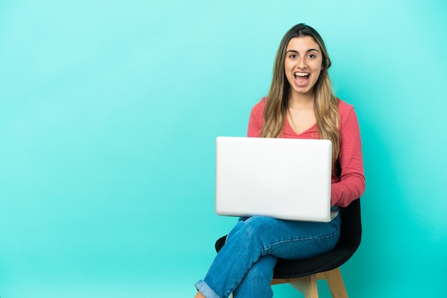 Jeune femme de race blanche assise sur une chaise avec son pc isolé sur fond bleu avec une expression faciale surprise