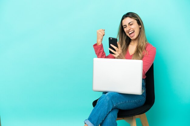 Jeune femme de race blanche assise sur une chaise avec son pc isolé sur fond bleu avec téléphone en position de victoire