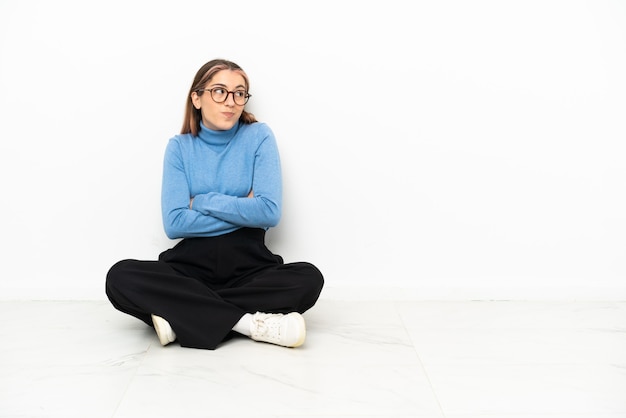 Jeune femme de race blanche assise sur le sol en faisant des doutes tout en soulevant les épaules