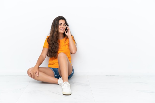 Jeune femme de race blanche assise sur le sol isolé sur fond blanc en gardant une conversation avec le téléphone portable avec quelqu'un