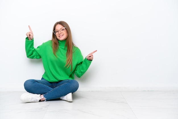 Jeune femme de race blanche assise sur le sol isolé sur fond blanc, pointant le doigt vers les latéraux et heureux