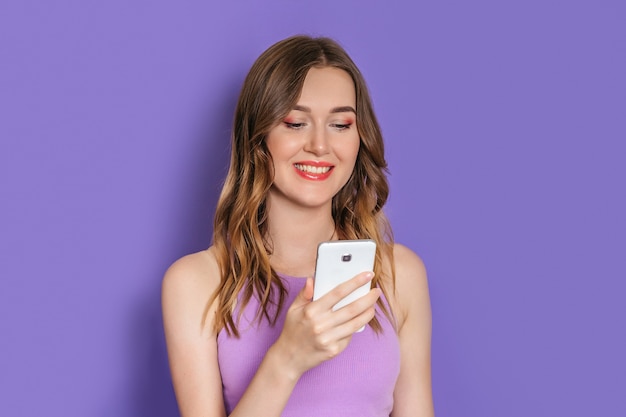 Jeune femme de race blanche aux cheveux bouclés souriant et regardant l'écran du téléphone portable isolé sur fond de studio lilas