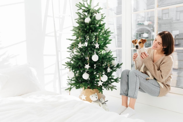 Une jeune femme de race blanche aux cheveux noirs avec un sourire agréable joue avec un chien de race porte un jean pull et des chaussettes est assise sur le rebord de la fenêtre près d'un lit blanc et d'un sapin de Noël décoré Vacances du Nouvel An