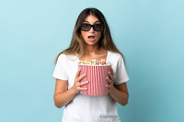 Jeune femme de race blanche sur bleu surpris avec des lunettes 3D et tenant un grand seau de pop-corn