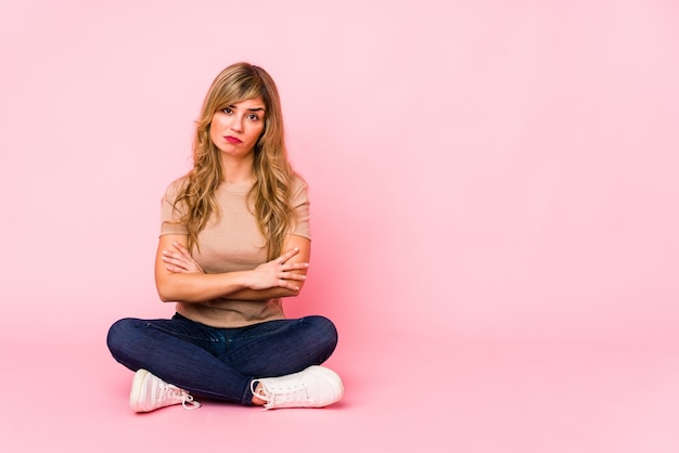 Jeune femme de race blanche blonde assise sur un studio rose malheureux à la recherche à huis clos avec une expression sarcastique.