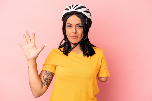 Jeune femme de race blanche avec un bras portant un casque de vélo isolé sur fond rose souriant joyeux montrant le numéro cinq avec les doigts.