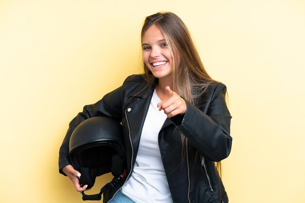 Jeune femme de race blanche avec un casque de moto isolé sur fond jaune pointant vers l'avant avec une expression heureuse