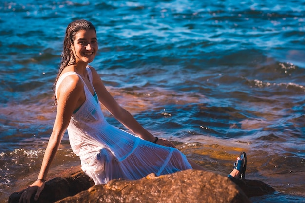 Une jeune femme de race blanche dans une robe blanche mouillée assise sur un rocher