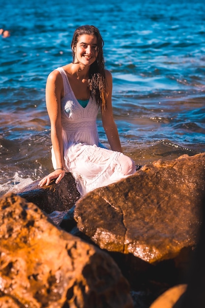 Une jeune femme de race blanche dans une robe blanche mouillée assise sur un rocher