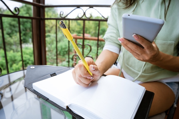 Jeune femme de race blanche écrit dans un cahier avec un stylo dans des vêtements décontractés avec un téléphone à la main sur un balcon d'été