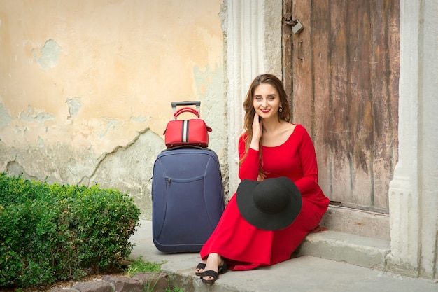 Une jeune femme de race blanche est assise sur le seuil d'une vieille maison dans une longue robe rouge avec valise à l'extérieur