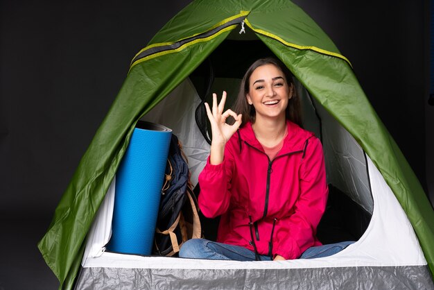 Jeune femme de race blanche à l'intérieur d'une tente de camping vert montrant signe ok avec les doigts