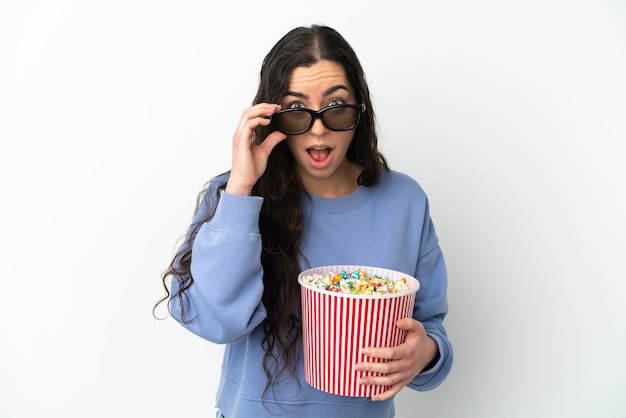 Jeune femme de race blanche isolée sur fond blanc surpris avec des lunettes 3D et tenant un grand seau de pop-corn