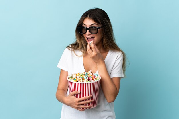 Jeune femme de race blanche isolée sur fond bleu avec des lunettes 3D et tenant un grand seau de pop-corn tout en regardant côté